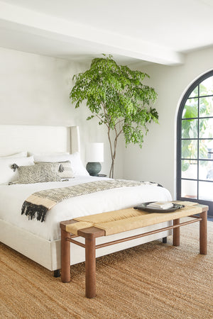 The Macie Jute Rug lays in a bright bedroom under a white linen framed bed and woven bench