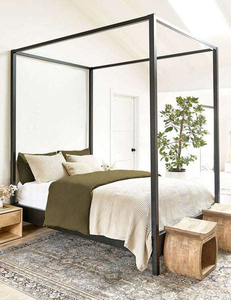 #color::olive #size::queen #size::king #size::twin #size::cal-king | The European Flax Linen olive green Duvet Set by Cultiver lays on a black canopy bed in a bedroom with cream waffle linens, a persian rug, and natural wooden ottomans and nightstands