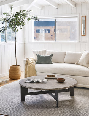 The Andra round coffee table with light mango wood top and iron X-base sits in a bright living room with wood paneled walls, a natural linen sofa, and a gray woven rug