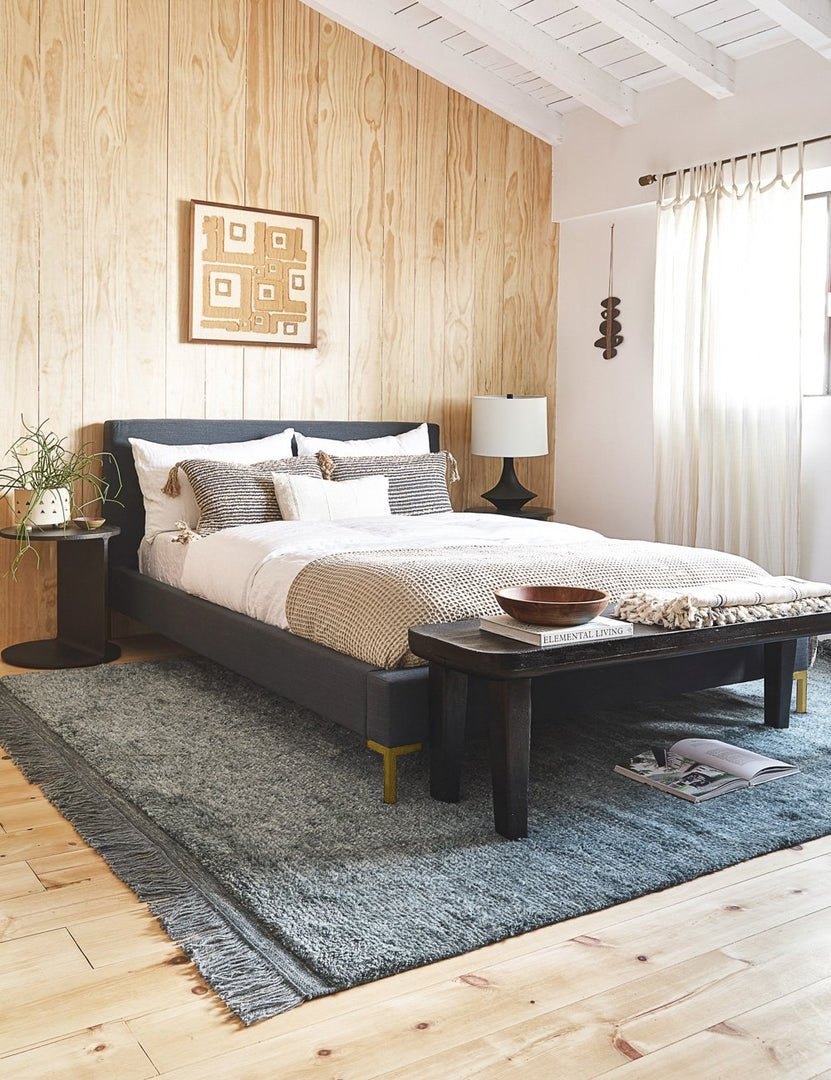 #color::navy-linen #size::twin #size::full #size::queen #size::king #size::cal-king | The Deva navy linen bed lays in a bedroom with a wooden paneled wall atop a gray fringed rug