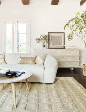 The Corliss 6-drawer white-washed wooden dresser sits behind a white curved sofa and round light wood coffee table atop a neutral tufted area rug.