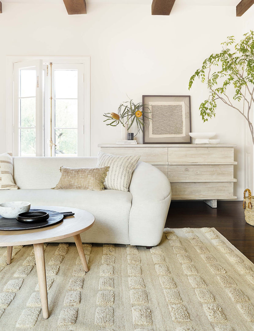 | The Corliss 6-drawer white-washed wooden dresser sits behind a white curved sofa and round light wood coffee table atop a neutral tufted area rug.