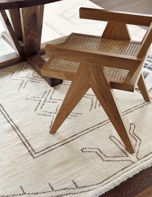The Rehya neutral geometric wool patterned rug sits in a dining room beneath a rattan dining chair and a wooden circular dining table with a sculptural base