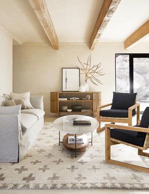 Two Regine wooden accent chairs with black cushions and rattan detailing sit in a living room across from a white oval coffee table and a light gray sofa, atop a plush patterned rug
