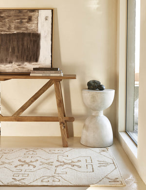 The Evet neutral geometric wool floor rug lays in a bright entryway in front of a wooden craftsman console table and round stone side table.