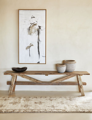 The Arlene craftsman-style antiqued teak wood bench sits against a wall with a black and white abstract painting above it, small vases on it, and a tan and ivory runner rug in front of it.