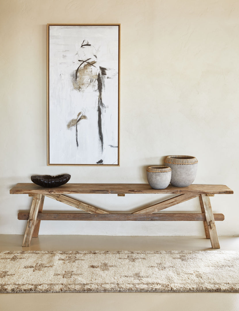 | The Arlene craftsman-style antiqued teak wood bench sits against a wall with a black and white abstract painting above it, small vases on it, and a tan and ivory runner rug in front of it.