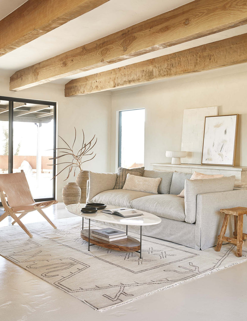 | The Thomas Bina oval coffee table with white laquered top, oak shelf and steel frame in a neutral living room with a light gray slipcovered sofa and light tan leather slingback chair under wooden ceiling beams.