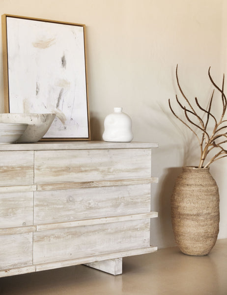 | The Corliss 6-drawer white-washed wooden dresser has a white abstract painting leaning on top of it and decorative white bowls and vases.