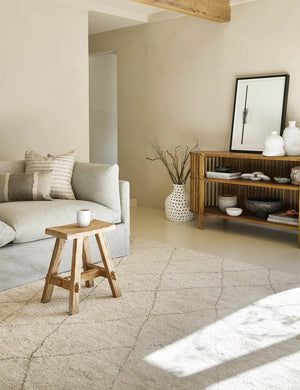 The afella rug lays in a living room with a textured wooden sideboard, a natural linen sofa, and a wooden stool