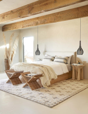 The Acoma cream and tan plus-sign patterned Moroccan area rug with diamond border lays under a bed with ivory upholstered headboard and wood frame with two wooden stools at the end of the bed.