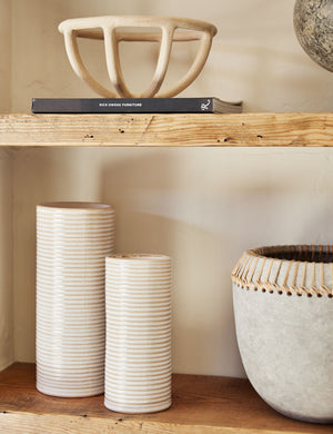 The Prong cream stoneware ceramic centerpiece bowl by SIN sits on a wooden shelf with narrow ribbed vases, a black paperback book, and a vase with jute detailing