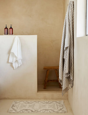 The Rehya neutral geometric wool patterned rug sits in a bathroom with a wooden stool, a gray and white striped throw, and a white tassel hand towel