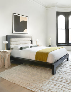 The Aimee mohair mustard yellow wool throw blanket with fringe ends lays at the edge of a black framed bed with a gray ribbed headboard in a bedroom with floor to ceiling black framed windows