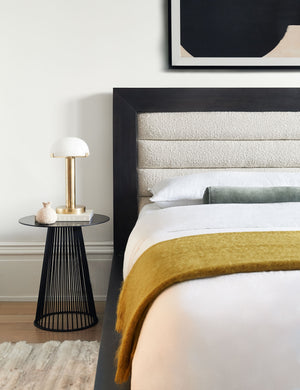 The Lelani table lamp with half-moon shade on a gold base sits in a bedroom atop a sculptural black nightstand to the left of a bed with a wooden framed boucle headboard