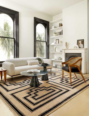 The Senna neutral hand-knotted wool area rug with black geometric pattern sits in a black and white living room with a white sofa, wood cane accent chair, and rounded black coffee tables.