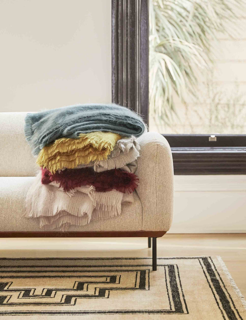 #color::blue | The Aimee mohair wool throw in mustard, blue, blush, warm gray, and merlot are stacked atop each other on a gray sofa in a living room with a geometric rug