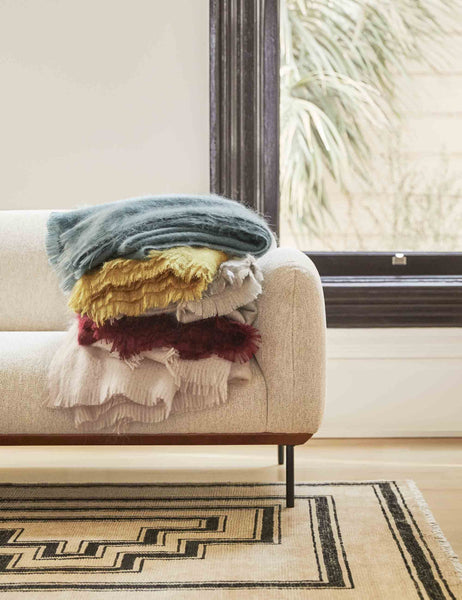 #color::blush | The Aimee mohair wool throw in mustard, blue, blush, warm gray, and merlot are stacked atop each other on a gray sofa in a living room with a geometric rug