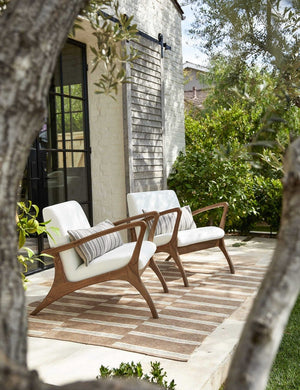 Two Venturi white indoor and outdoor accent chairs with wooden legs sit on a patio outside a white brick house atop a brown and white striped rug.