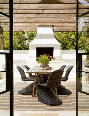The Manila wicker weave black indoor and outdoor dining chair sits in an outdoor dining space underneath a wood paneled covering with a large white fireplace in the background.