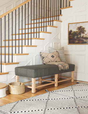 The ambleside loden gray linen bench sits by a staircase against white accented walls near a black and white patterned rug