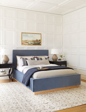 The Lockwood blue velvet-upholstered bed with a white oak base sits atop a patterned hand-woven rug against a white, accented wall.