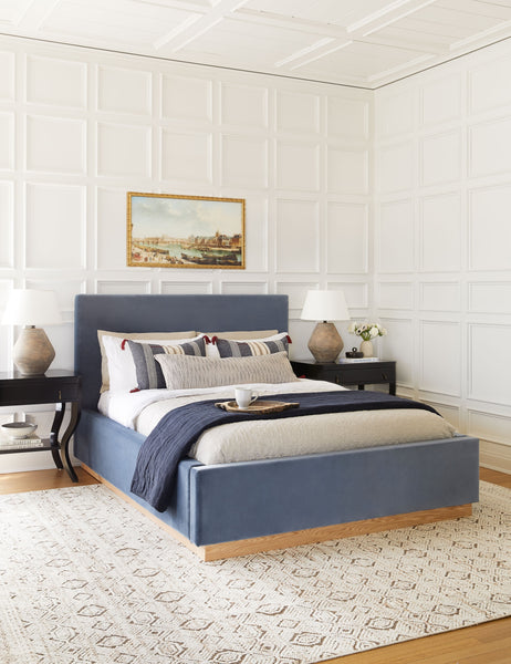 #color::harbor #size::cal-king #size::king #size::queen | The Lockwood blue velvet-upholstered bed with a white oak base sits atop a patterned hand-woven rug against a white, accented wall.