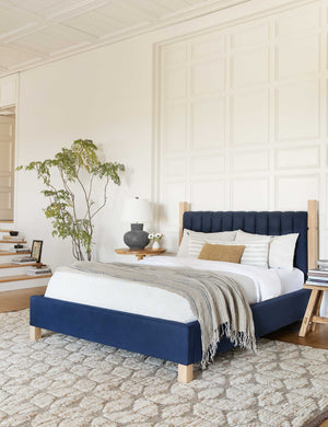 The Ambleside dark blue velvet bed sits atop a patterned rug in between two circular wooden nightstands