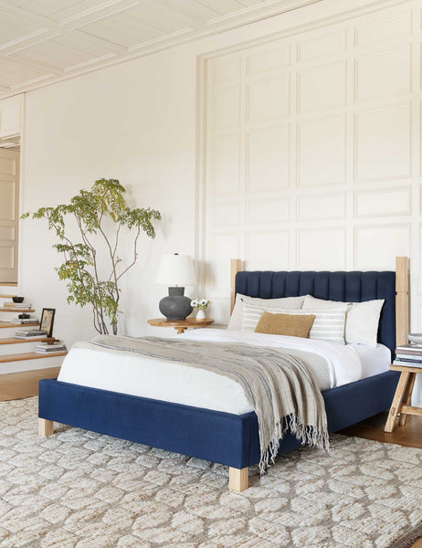 #size::cal-king #size::king #color::dark-blue #size::queen | The Ambleside dark blue velvet bed sits atop a patterned rug in between two circular wooden nightstands 