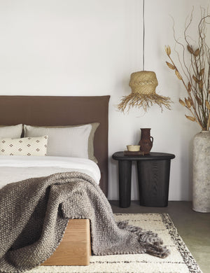 The Olema mink gray handwoven throw with fringed ends lays in a bedroom with a cream and black plush rug, a wooden-base bed, and a black sculptural nightstand