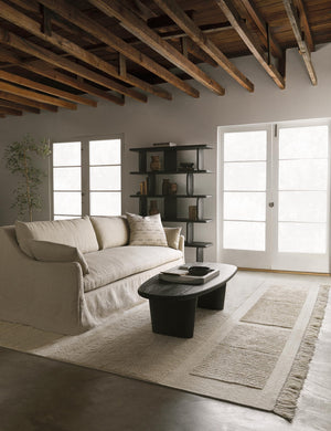 The Portola Natural linen Slipcover Sofa sits atop an ivory textured rug next to a black coffee table and bookshelf