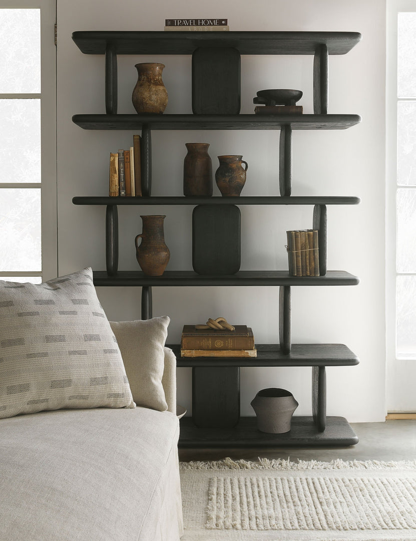 #color::black | The Nera black solid wood sculptural bookcase sits in a living room with an ivory couch and rug, patterned throw pillow, and various books and vases stacked within its shelving.