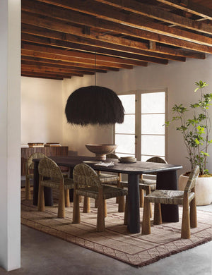 The Paige black plush pendant light with palm tree fibers sits in a dining room with wooden beamed walls, jute dining chairs, and a brown patterned rug