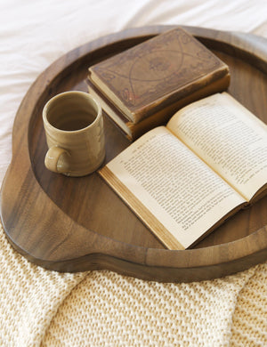 The Lylah round wooden ochre tray sits atop a textured cream throw pillow with a ceramic mug and two books