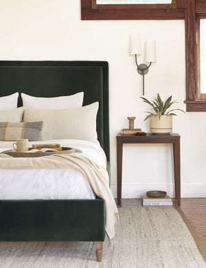The Sassa old bronze asymmetrical double sconce with white-shades is mounted in a bedroom with a emerald velvet framed bed, a wooden nightstand, and a natural-toned woven rug
