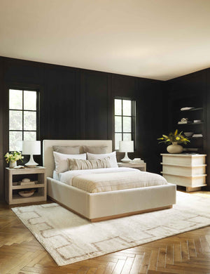 The Lockwood natural velvet-upholstered bed with a white oak base sits in a monochrome bedroom atop a fluffy patterned rug against dark paneled walls.