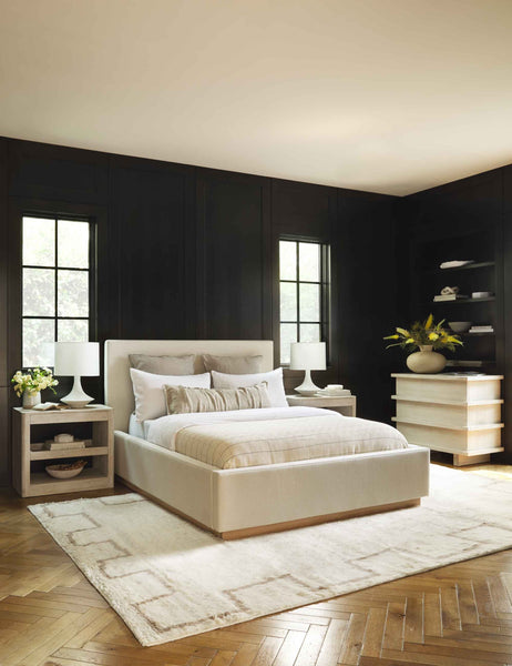#color::natural #size::cal-king #size::king #size::queen  | The Lockwood natural velvet-upholstered bed with a white oak base sits in a monochrome bedroom atop a fluffy patterned rug against dark paneled walls.