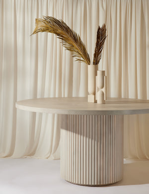 The Rutherford white-washed acacia wood round dining table sits below two white sculptural vases with white curtains draped behind it.