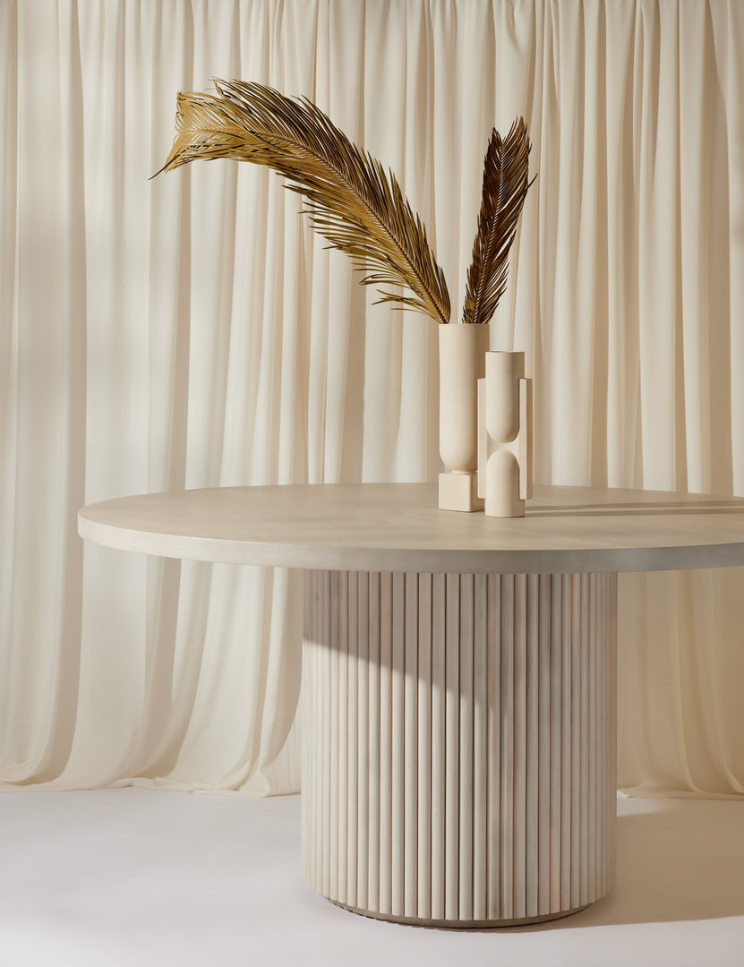 | The Rutherford white-washed acacia wood round dining table sits below two white sculptural vases with white curtains draped behind it.