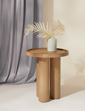 The Delta natural wooden side table with pedestal base sits in a room with a white cylindrical vase atop it and a gray curtain draped behind it