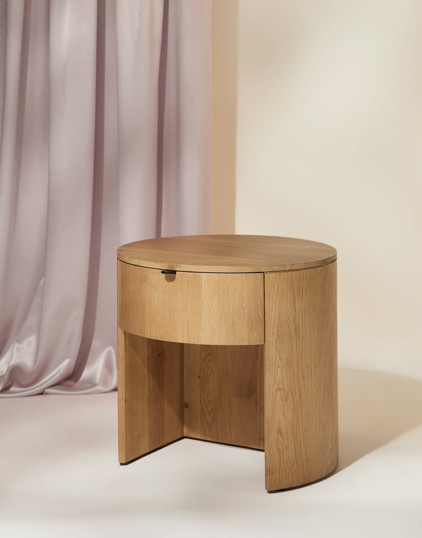 | Angled right view of the Kono round solid oak nightstand with one drawer on a purple draped backdrop