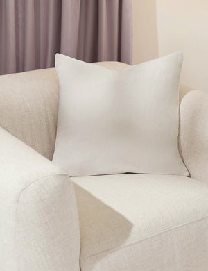 Charlotte oyster white square velvet pillow sits on a white accent chair with a purple curtain in the background