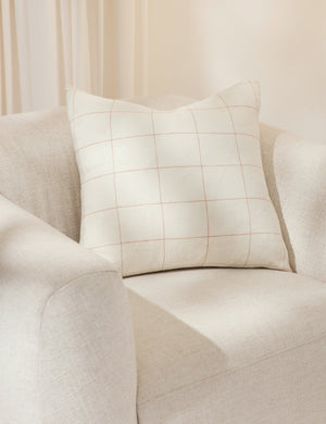The lucian white and rust square pillow sits atop an ivory accent chair in a studio room with white curtains