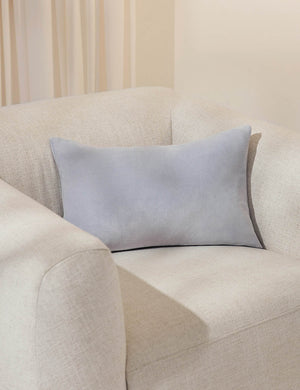 Charlotte ice blue lumbar velvet pillow sits on a white accent chair with a white curtain in the background