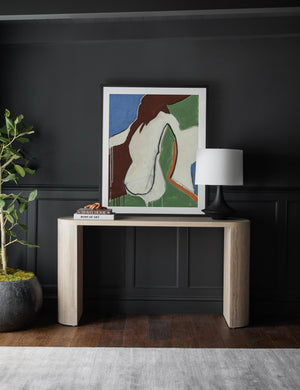The Luna white-washed oak oval console table sits against a black wall with a multicolored abstract wall art, stack of books, and black sculptural lamp sitting atop it.