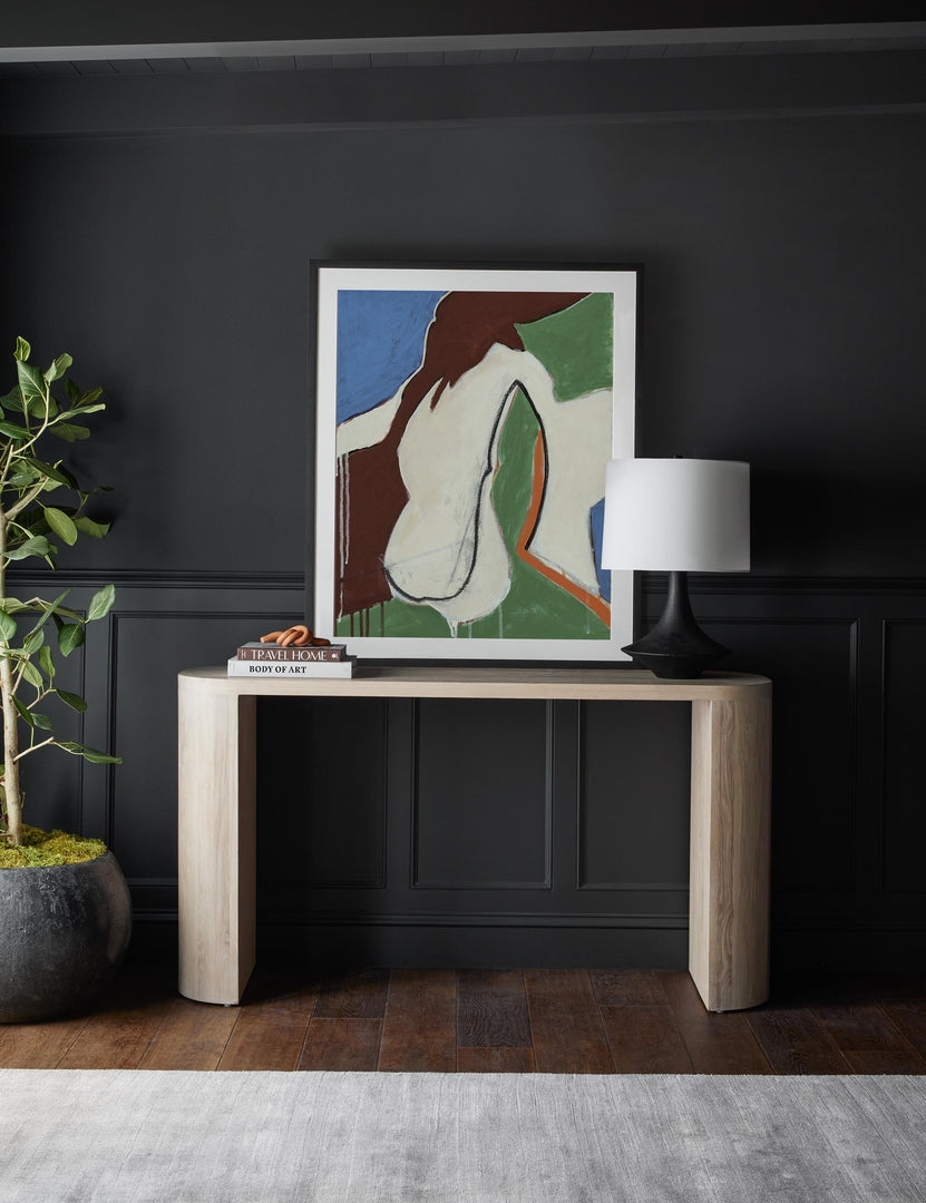 #color::Washed-Oak | The Luna white-washed oak oval console table sits against a black wall with a multicolored abstract wall art, stack of books, and black sculptural lamp sitting atop it.