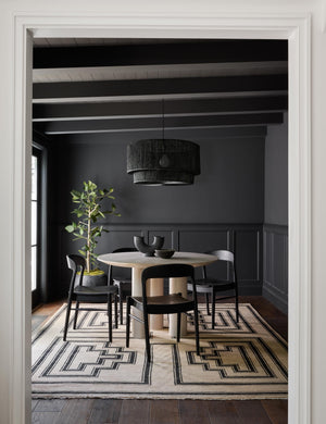 The Senna neutral hand-knotted wool area rug with black geometric pattern sits in a black dining room with a round white dining table, four black wooden dining chairs, and black chandelier
