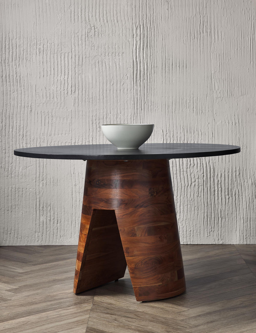 | The Adler acadia wooden round dining table sits in a studio with a white bowl atop chevron floors in front of a textured wall