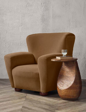 The Avery Bronze Mohair accent chair sits in a studio next to a sculptural textured side table with a glass of water and book