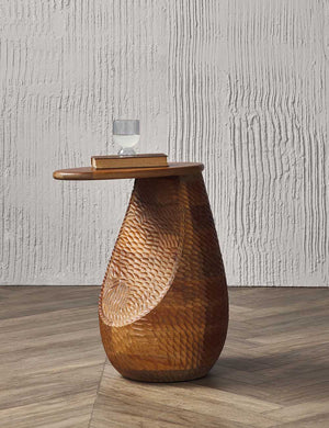 The Gem mango wood side table sits in a studio with chevron wooden floors and textured walls with a book and glass atop it